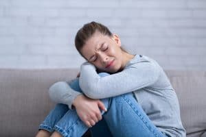 Miscarriage Support. Young depressed woman sitting on sofa with closed eyes and hugging her knees, feeling desperate and stressed.