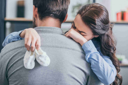woman crying, hugging man and holding baby socks at home, grieving disorder concept