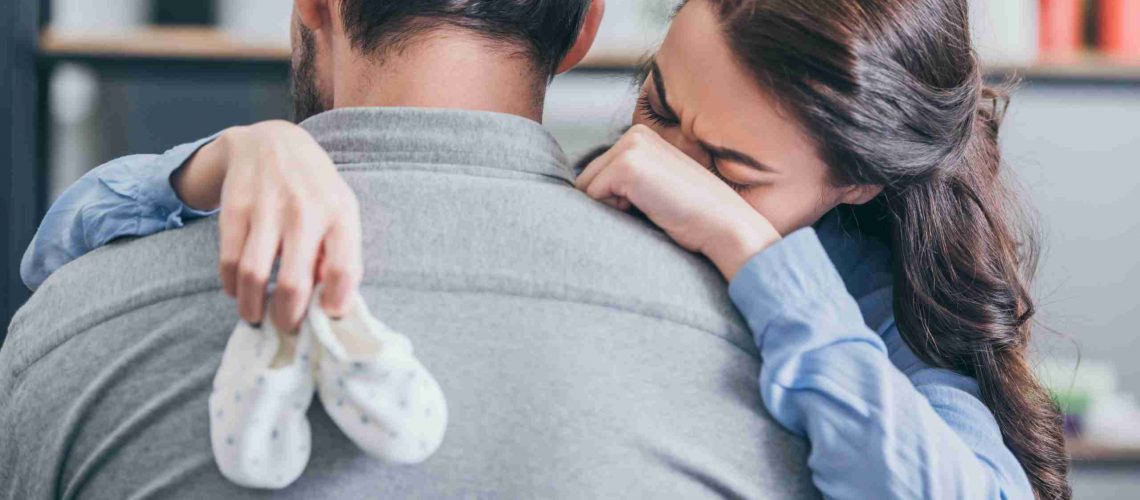 woman crying, hugging man and holding baby socks at home, grieving disorder concept