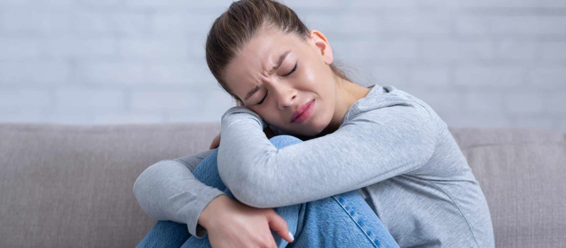 Miscarriage Support. Young depressed woman sitting on sofa with closed eyes and hugging her knees, feeling desperate and stressed.
